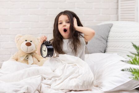 cute surprised little child girl in pyjamas with clock in bed. good morningの写真素材