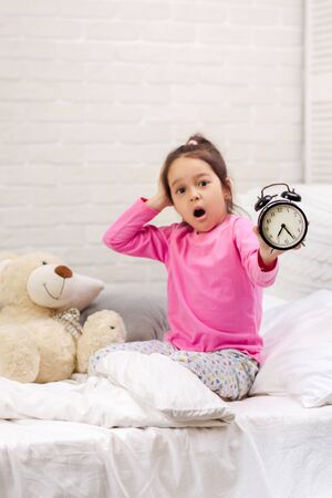 Surprised cute little child girl in pyjamas with clock in bed. good morningの写真素材