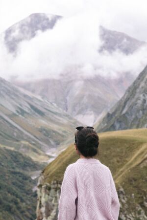 young girl in a sweater is looking at the Caucasian mountains, Georgia, rear view.