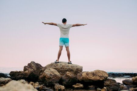 Young man standing with raised hands on top of a mountain sea and enjoying view. man enjoying the sensation of freedom in front of setting sunの写真素材