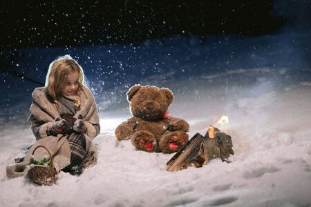 cute little girl sitting and warming herself by the fire in the winter evening. fantastic night by the fire. frame as illustration to a fairy taleの写真素材