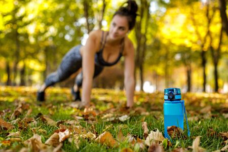 blue bottle of water on green grass. sporty woman doing outdoor workout in park on background. focus on bottleの写真素材
