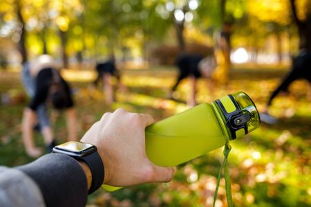 male hand holding bottle of water on background of sporty women doing outdoor workout in park. focus on bottle. helthy lifestyleの写真素材