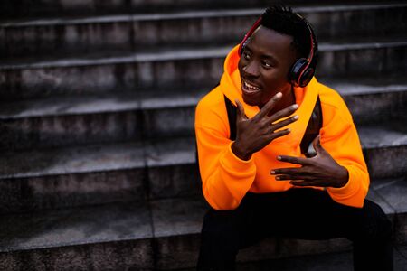 smiling african-american man in stylish orange hoodie sweatshirt in wireless headphones listening music and sitting on stairs. copy spaceの写真素材