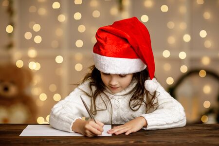Happy little child girl in santa hat writing wish list or letter to Santa Claus on background with lights. Merry Christmasの写真素材