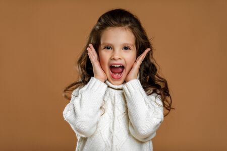 happy surprised beautiful curly little child girl in white sweater looking to camera on beige...
