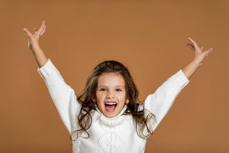Cute happy smiling little child girl in white sweater raising her hands up on beige background.