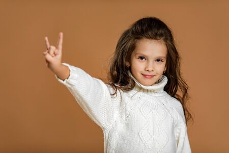 Cute smiling little child girl in white sweater making Rock gesture on beige background.の写真素材
