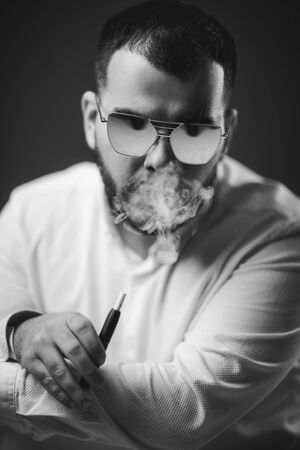 Portrait of bearded man in sunglasses and white shirt smoking on black background. Vaping man.
