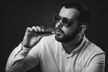 Portrait of bearded man in sunglasses and white shirt smoking on black background. Vaping man.の写真素材