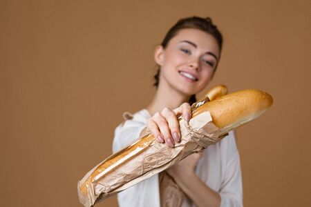 beautiful smiling young woman holding bread in paper bag on studio yellow background. girl showing fresh fragrant long loaf. focus on breadの写真素材
