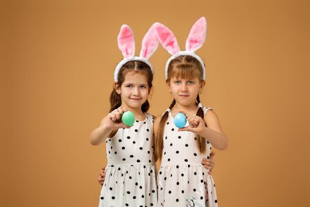 happy cute little child girls with pink bunny ears holding painted Easter eggs on studio yellow...