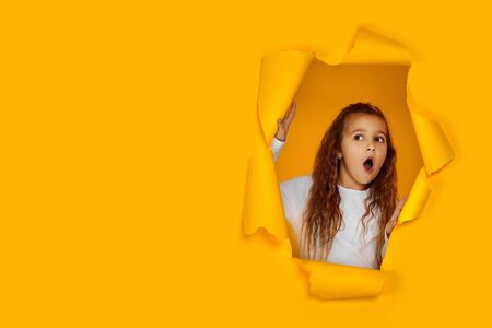 Portrait of cute surprised caucasian little child girl looking with happy face expression at the camera in torn paper yellow wall. copy space. banner for discounts and salesの写真素材