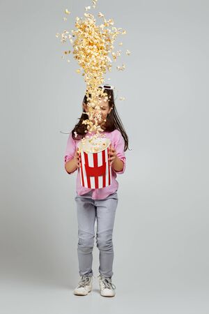 full length view of smiling caucasian child girl in red-blue 3d glasses holding bucket with flying popcorn and jumping on gray background.の写真素材