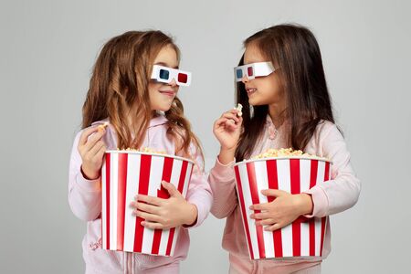 two beautiful caucasian little girls wearing red-blue 3d glasses and eating popcorn from bucket on...
