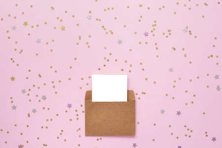 white blank card in kraft brown paper envelope on pink background with golden confetti. Holiday...