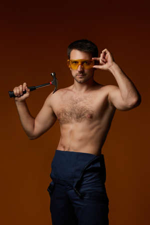 sexy caucasian man worker in overalls holding hammer isolated on orange studio backgroundの写真素材