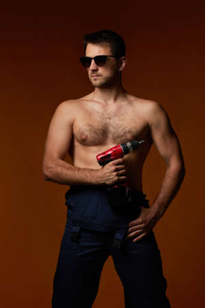 sexy caucasian man in worker overalls holding cordless screwdriver isolated on brown studio backgroundの写真素材