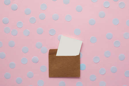 white blank card in kraft brown paper envelope on pink background with confetti. Holiday mockup. Top...