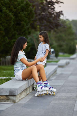 happy little child girl and her mother are wearing roller skates while sitting in summer park.