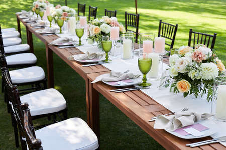 chairs and dinner tables with white cloth, served with porcelain and green glasses. Georgeous event party table decorated with flowers on a green lawnの写真素材