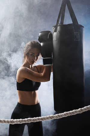 athletic woman in black boxing gloves leaning on punching bag on dark studio background with smoke