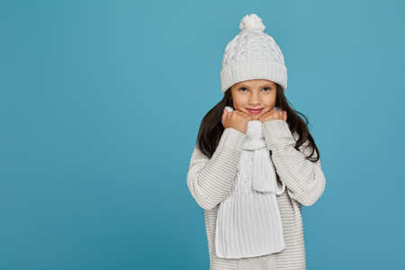 portrait of smiling cheerful child girl in white winter knitted hat and sweater isolated on blue...