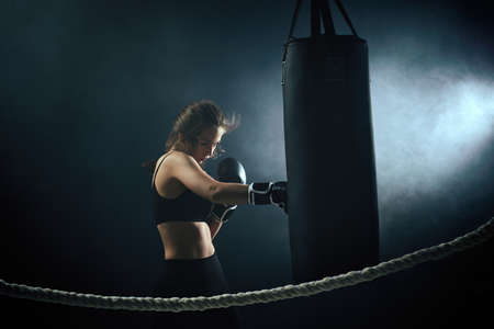 sporty brunette woman in boxing gloves and sportswear hitting the boxing bag in dark background with smoke.の写真素材