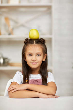 little child girl sitting at the table with apple on head in the kitchen.の写真素材