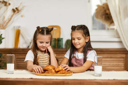 happy little children girls having breakfast - fresh delicious croissants and milk in the kitchen.の写真素材