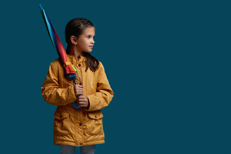 cute child girl with multicolored umbrella in yellow rain coat on blue background. autumn mood. copy...