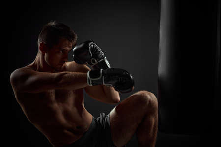 muscular boxer in black boxing gloves punching in boxing bag on dark background