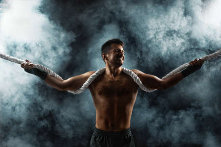 strong athletic muscular man with naked body holding rope on smoke backgroundの写真素材