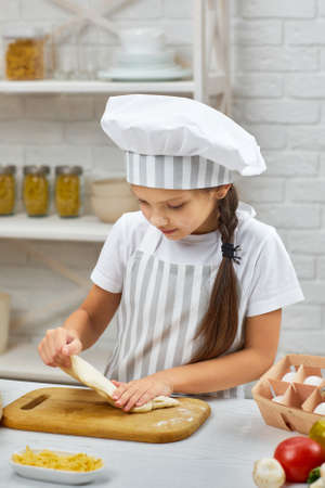 little child girl in cap and an apron knead the dough in the kitchen