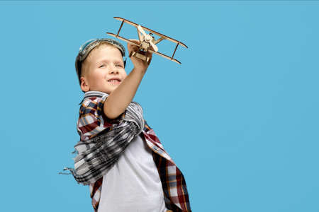happy child boy in retro glasses and scarf playing with wooden toy airplane on blue background....