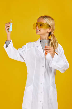little child girl in protective glasses holding a chemical flask and test tube on yellow studio background. Choice of profession. Doctor, laboratory assistant, chemist.の写真素材