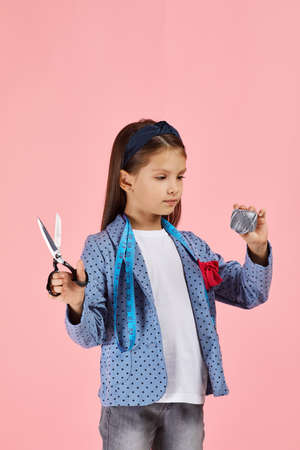 adorable little girl with measuring tape holding scissors and spool of thread on pink background. the child dreams of becoming a tailor or fashion designerの写真素材