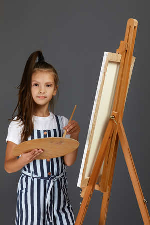 Portrait of beautiful little girl holding a wooden art palette and brush on gray studio background....