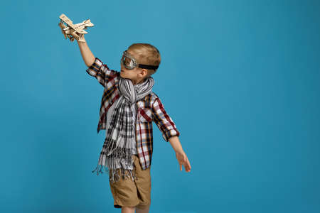 happy child boy in retro glasses and scarf playing with wooden toy airplane on blue background....