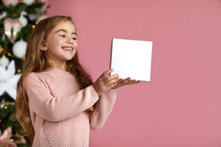 beautiful happy little child girl holding Christmas gift box in hand over the pink background and christmas tree. copy space for textの写真素材