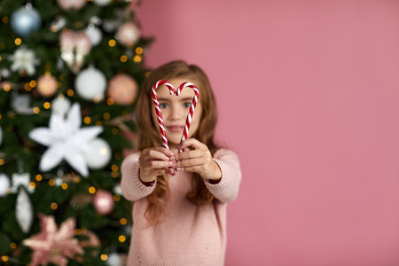 cute little child girl holding heart-shaped lollipop cane on the pink background of christmas tree. copy space. focus on candyの写真素材