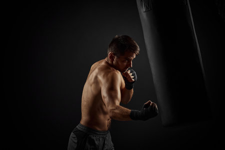 Aggressive shirtless boxer punching in boxing bag on black backgroundの写真素材