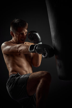muscular boxer in black boxing gloves punching in boxing bag on dark backgroundの写真素材