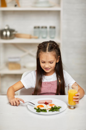 little child girl having breakfast - fried egg and orange juice in the kitchen. healthy breakfast. fried egg face smiles on a white plateの写真素材