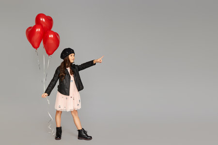 stylish child girl holding red heart shaped balloons and pointing to the side isolated on gray background. valentines day. copy spaceの写真素材