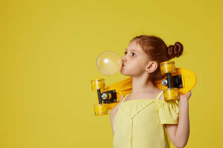 Stylish little child girl with yellow skateboard chewing gum over yellow background