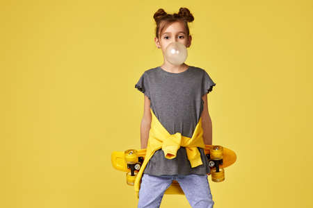 happy little child girl with yellow skateboard blowing a big bubble gum over yellow background. copy...