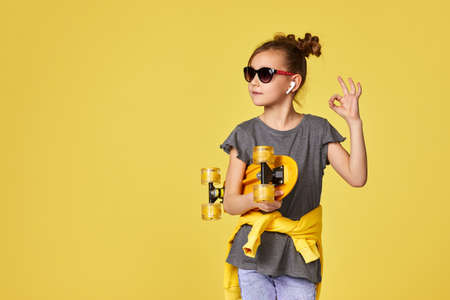 Stylish cool little child girl in sunglasses with yellow skateboard showing Ok gesture on yellow...