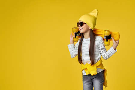 smiling little child girl in hat and sunglasses posing with yellow skateboard on yellow background. copy spaceの写真素材