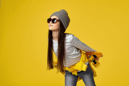 cool little child girl in hat and sunglasses posing with yellow skateboard on yellow background.の写真素材